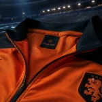 Veste pays bas orange/noir 2026, vue entraînement sur banc, stade flou